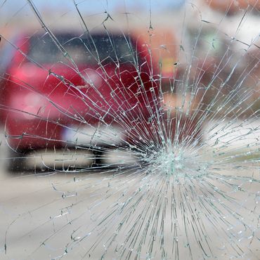 A shattered car windshield shot from inside, with a red car and a blurred street scene visible in the background. The cracks form a circular pattern, radiating outward from a central point.