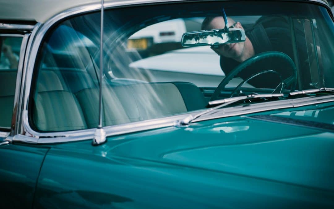 A person leans against the open driver’s side window of a vintage green car with a beige roof, peering inside. The interior appears well-maintained, and the car's reflections gleam on the windshield—an excellent example of diligent car maintenance.