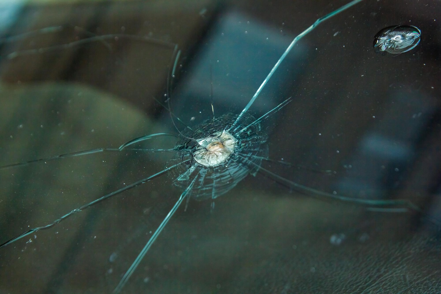 Close-up of a car windshield showcasing a large circular crack with radiating lines, hinting at impact damage. A small chip is visible nearby. The blurred background suggests a vehicle interior, highlighting the importance of cracking prevention for car windows.