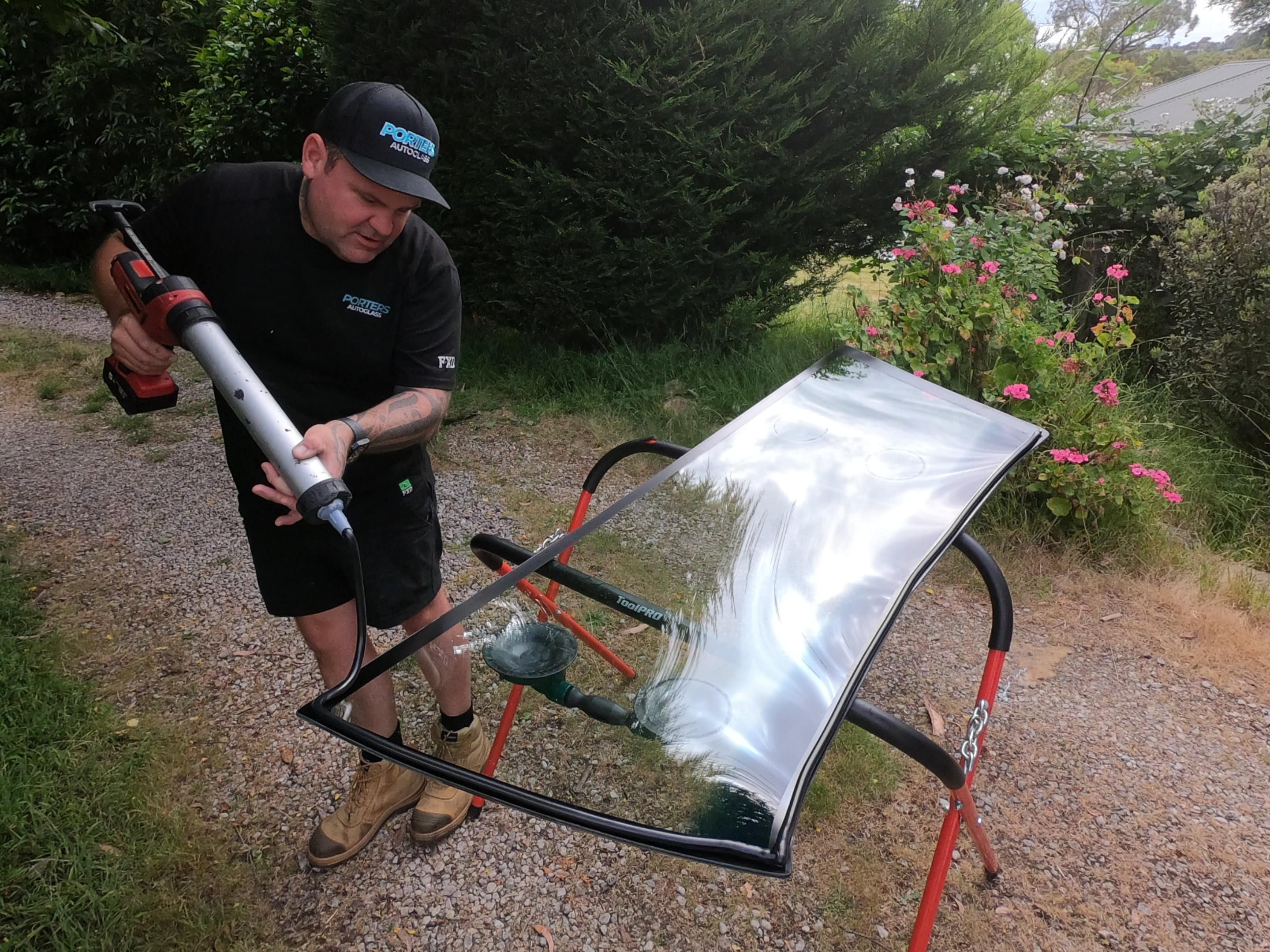 A person in a black shirt and cap is applying adhesive to the edge of a windshield set on a stand outdoors They are using a caulking gun and are surrounded by greenery and flowers