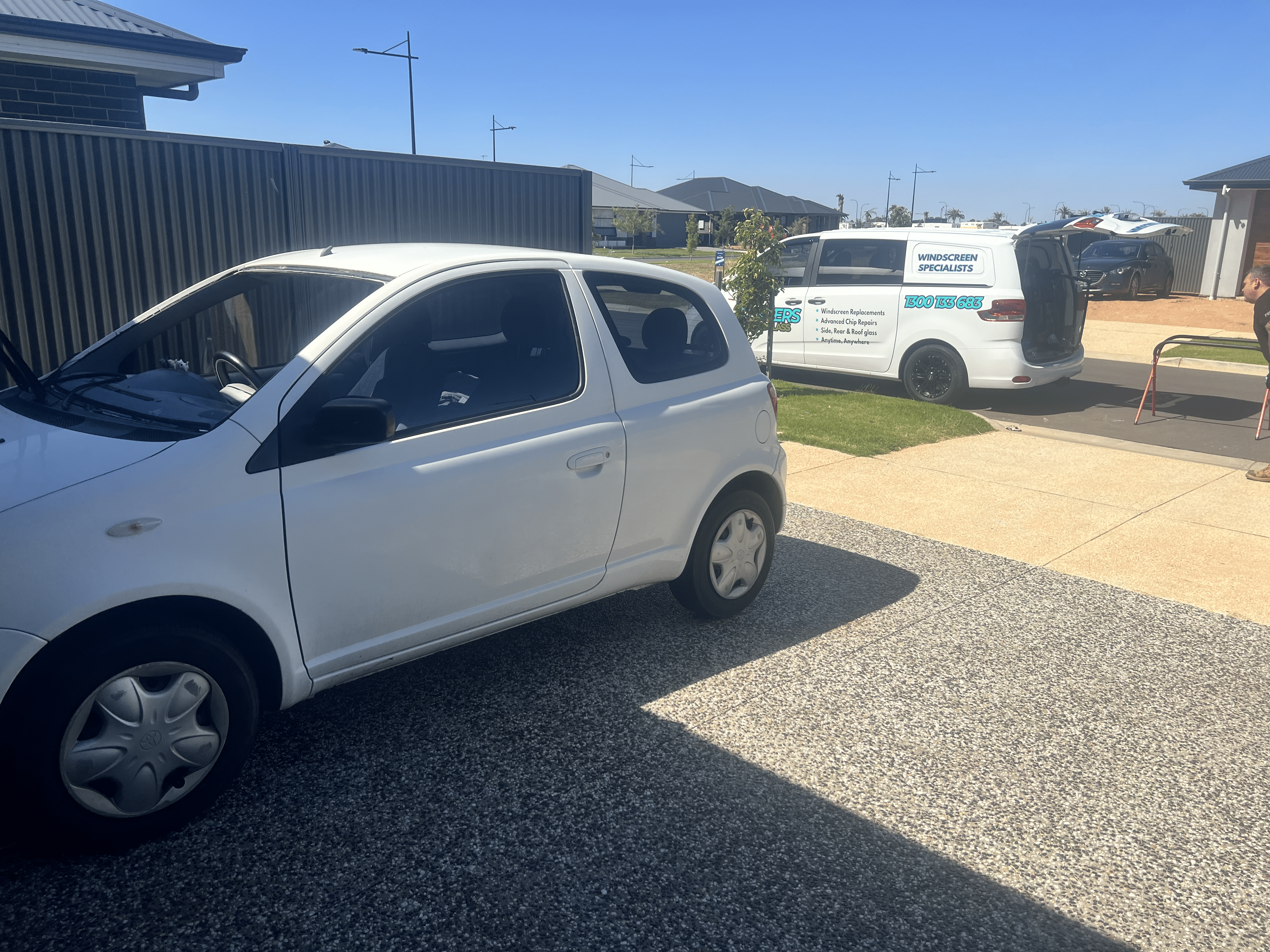 A white compact car is parked in a driveway on a sunny day In the background, a van labeled "Windscreen Specialists" is parked on the street Residential houses and a clear sky are visible